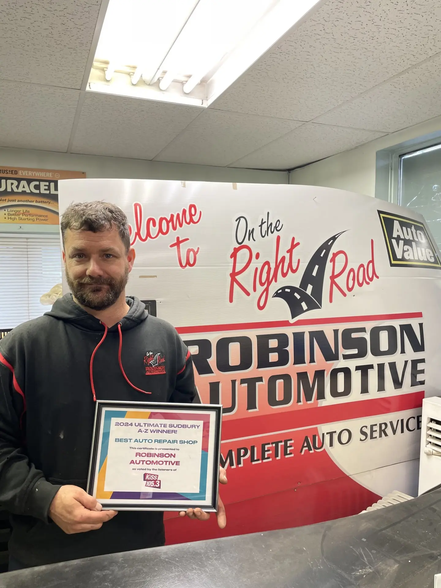 Robinson Automotive owner holding the 2024 Ultimate Sudbury A–Z award for Best Auto Repair Shop, in front of the On the Right Road shop banner